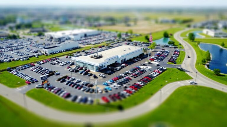 Aerial high-angle view of a bustling car dealership surrounded by parked cars in a green landscape.